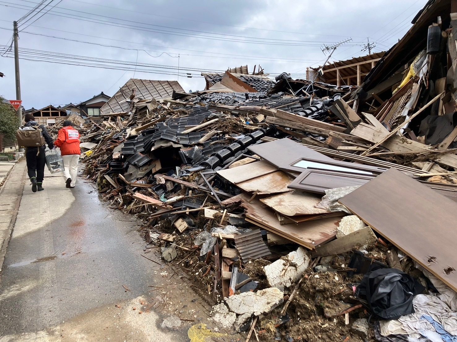 能登半島地震の被災地で、瓦礫のそばをキャリーを持った人とオレンジのベストの支援者が歩く